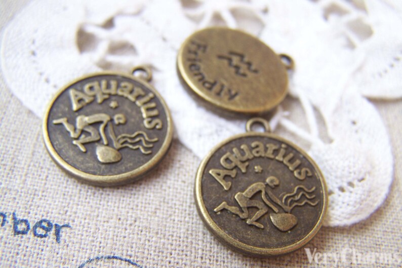 May include: Three bronze-colored metal charms with the zodiac sign Aquarius. The charms are round and have a raised design of the Aquarius symbol and the word "Aquarius" written around the edge. One charm has the word "Friendly" written on the back.