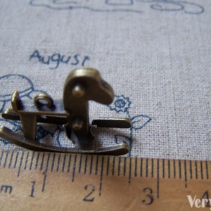 Puede incluir: Un peque&ntilde;o amuleto de caballo de balanc&iacute;n de color dorado antiguo, que mide aproximadamente 2 cm de largo. El amuleto est&aacute; detallado con una base de balanc&iacute;n y una cabeza de caballo con una crin levantada.