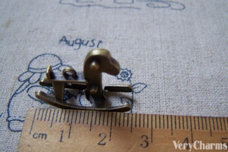 Puede incluir: Un peque&ntilde;o amuleto de caballo de balanc&iacute;n de color dorado antiguo, que mide aproximadamente 2 cm de largo. El amuleto est&aacute; detallado con una base de balanc&iacute;n y una cabeza de caballo con una crin levantada.
