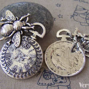 May include: Two antique silver-toned charms featuring a bee perched on a pocket watch. One charm has a clock face with Roman numerals, the other has a blank face.