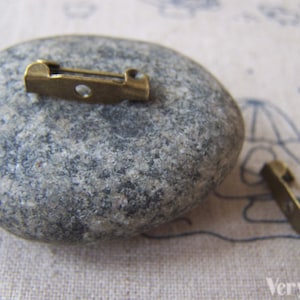 May include: Two antique gold-toned metal safety pin closures. The pins are on a gray stone surface.