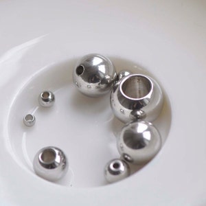 May include: A collection of silver metal beads of various sizes in a white bowl. The beads are smooth and shiny.