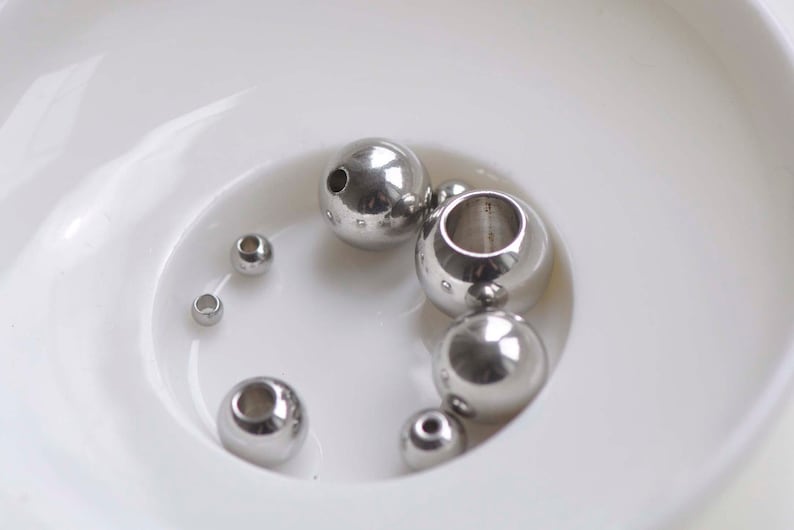 May include: A collection of silver metal beads of various sizes in a white bowl. The beads are smooth and shiny.