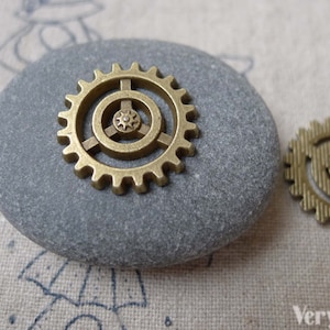 May include: Two gold-toned metal gears, one with a smaller gear inside, on a gray stone. The larger gear has a detailed design with multiple spokes and teeth.