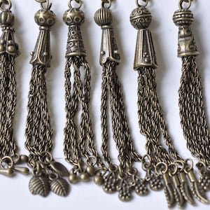 May include: An assortment of antique bronze-colored tassels. Each tassel features a decorative top with intricate designs and a chain of smaller chains. Some tassels have leaf and bead charms.