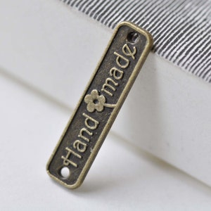 May include: A small, rectangular, bronze-colored metal tag with a flower design and the word "Handmade" engraved on it.