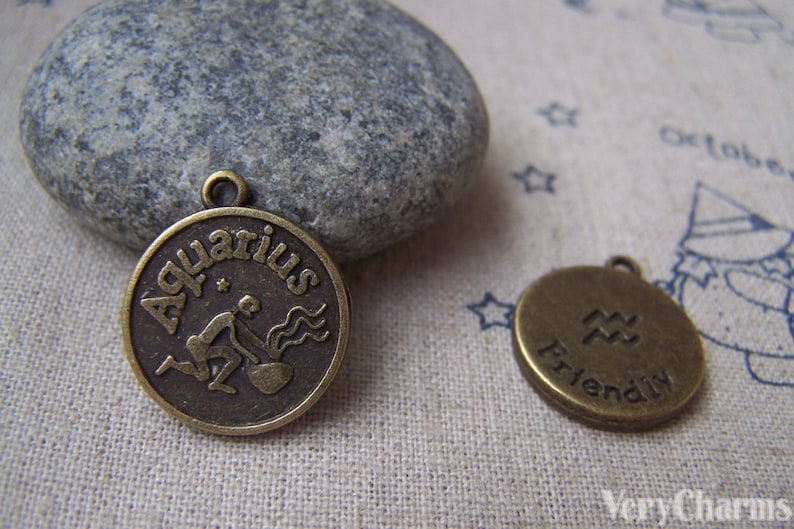 May include: Two bronze-colored metal charms with an Aquarius zodiac sign. The first charm features the word "Aquarius" written around the zodiac symbol. The second charm features the zodiac symbol and the word "Friendly".