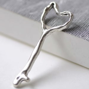 May include: A silver key charm with a heart-shaped loop at the top. The key is textured and has a rustic, antique look.