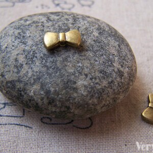 Op de afbeelding: Twee gouden strikcharms op een grijze stenen achtergrond. De charms zijn klein en delicaat, met een eenvoudig ontwerp.