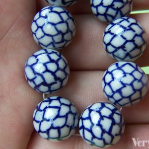 Op de afbeelding: Een streng witte keramische kralen met een blauw geometrisch patroon. De kralen zijn rond en aan elkaar geregen, met een gedetailleerd ontwerp. De kralen hebben een diameter van ongeveer 1,5 cm.