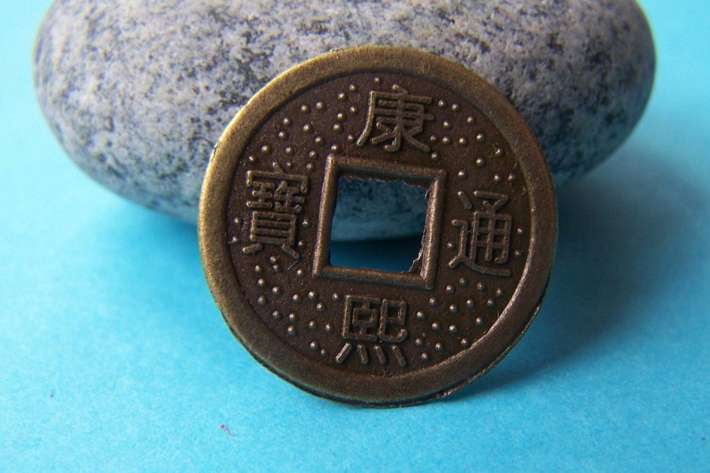 K&ouml;nnte beinhalten: Eine bronzene chinesische M&uuml;nze mit einem quadratischen Loch in der Mitte. Die M&uuml;nze tr&auml;gt chinesische Schriftzeichen, darunter "Wohlstand" und "Gl&uuml;ck".