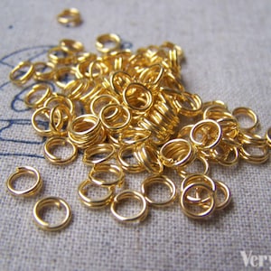 Op de afbeelding: Een grote stapel gouden springringen, een soort sieraadbevestiging, op een getextureerd stofoppervlak. De ringen zijn klein en rond en perfect om verschillende sieraden aan elkaar te verbinden.