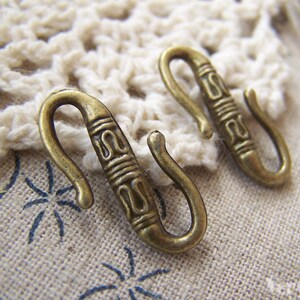 May include: Two antique gold-toned metal hooks with decorative designs. The hooks are S-shaped and have a textured surface.