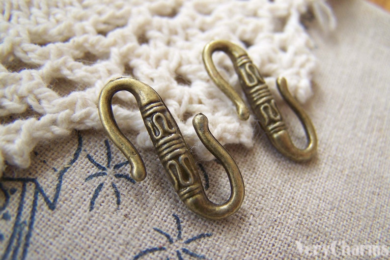 May include: Two antique gold-toned metal hooks with decorative designs. The hooks are S-shaped and have a textured surface.