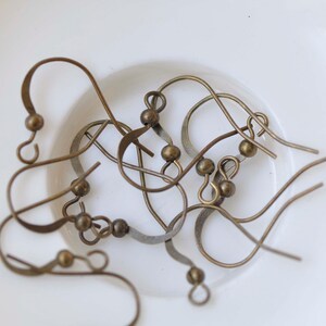 May include: A close-up of a group of gold-toned earring hooks. The hooks are made of metal and have a small loop at the top. The hooks are lying on a white surface.