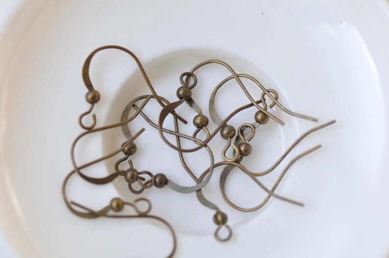 May include: A close-up of a group of gold-toned earring hooks. The hooks are made of metal and have a small loop at the top. The hooks are lying on a white surface.