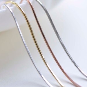 Könnte beinhalten: Drei silberne Ketten in verschiedenen Ausführungen: Silber, Gold und Roségold. Die Ketten sind auf einer weißen Oberfläche in einer Kurve angeordnet.