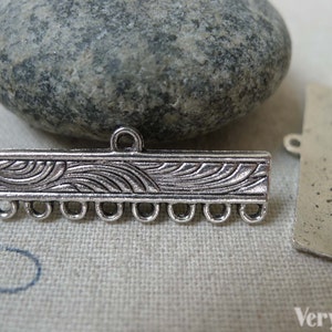 May include: Silver-toned metal connector with a decorative wave pattern and multiple loops for attaching jewellery or other crafts. The connector is shown on a fabric background with a stone.