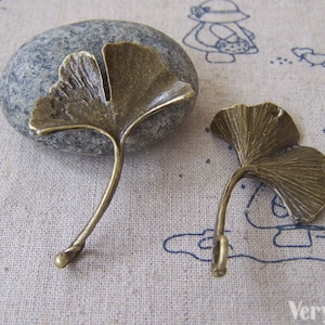 May include: Two antique bronze ginkgo leaf charms with a detailed design. The leaves are attached to a stem that curves into a loop.