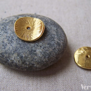 May include: Two gold-coloured metal round disc charms with a textured surface and a small hole in the centre. The charms are on a grey stone background.