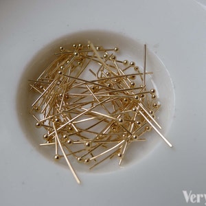 May include: A close-up of a white bowl filled with a large quantity of gold-colored head pins. The pins are approximately 1 inch long and have a small round head.