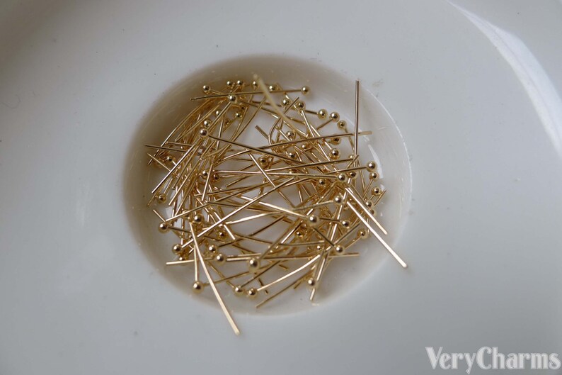 May include: A close-up of a white bowl filled with a large quantity of gold-colored head pins. The pins are approximately 1 inch long and have a small round head.