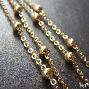 May include: Close-up of multiple strands of delicate gold necklaces. Each chain features small, round gold beads interspersed along the length. The chains are displayed against a dark, textured background, highlighting the jewelry's shine.