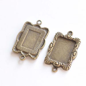 May include: Two antique bronze-colored rectangular pendant trays with ornate, scalloped edges. Each has a small loop at the top and bottom for attaching to a chain or other jewelry component. The trays are empty, ready for resin or other fillings.