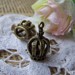 May include: Two antique gold-toned crown charms with intricate details. The charms are on a white lace background.