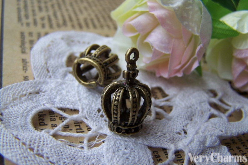 May include: Two antique gold-toned crown charms with intricate details. The charms are on a white lace background.