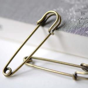 May include: Two antique gold safety pins, one lying on top of the other. The pins are made of metal and have a shiny finish.