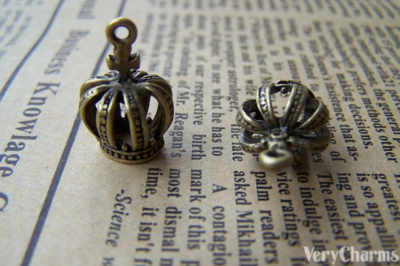 May include: Two antique gold-toned crown charms with a detailed design. The crowns have a pointed top and a loop for attaching to a necklace or bracelet.