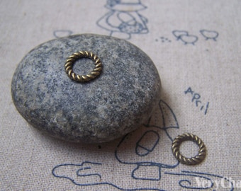 May include: Two small, gold-colored, metal jump rings. The rings are round and have a rope-like texture. They are sitting on a gray rock and a beige fabric surface.