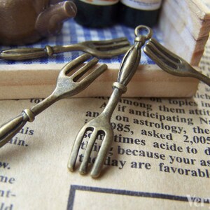 May include: Four antique bronze fork charms with a loop at the top for attaching to a necklace or bracelet. The forks are detailed with a classic design and have a rustic finish.