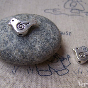 Könnte beinhalten: Zwei silberne Vogel-Anhänger aus Metall mit einem Wirbel-Design. Die Anhänger liegen auf einem grauen Stein und einem beigefarbenen Stoffhintergrund.