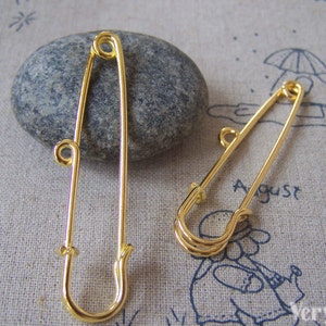 May include: Two gold safety pins, one larger than the other, are displayed on a textured beige surface. The larger pin is on a grey rock. The smaller pin is on the surface with a drawing of an umbrella and the word "August".