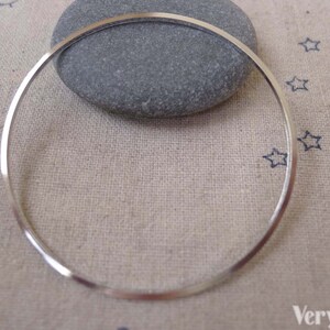 Può includere: Un braccialetto rigido in argento con una finitura liscia e lucida. Il braccialetto è appoggiato su una superficie di tessuto beige con un piccolo sasso e un disegno di una stella.