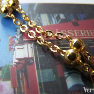May include: Close-up of a gold-colored necklace with a delicate chain and small, round beads. The necklace is draped over a postcard with a blurred image of a cafe and street scene.
