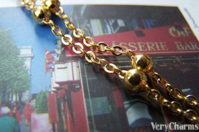 May include: Close-up of a gold-colored necklace with a delicate chain and small, round beads. The necklace is draped over a postcard with a blurred image of a cafe and street scene.