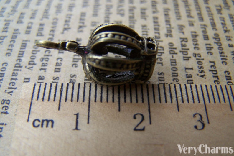 May include: A small, gold-colored crown charm with a single, clear rhinestone. The charm is about 1.5 cm long and is shown next to a ruler.