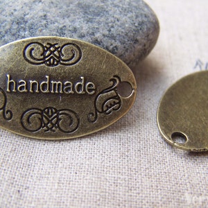May include: Two oval metal tags, one with the word "handmade" engraved on it and the other blank. Both tags have two holes for attaching to a project.