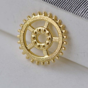 May include: Gold-colored metal gear with a detailed design. The gear has a circular shape with a smaller circle in the center. The gear has a textured surface.