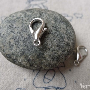 May include: Two silver lobster claw clasps, a type of jewelry finding, are shown on a gray stone background.