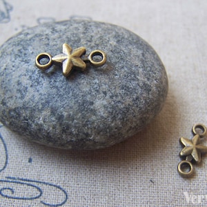 Puede incluir: Dos abalorios conectores en forma de estrella de bronce antiguo con anillas de salto. Los abalorios están sobre una superficie de piedra gris.