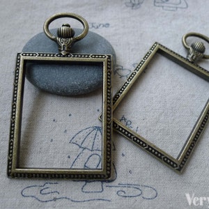 Könnte beinhalten: Zwei antike goldfarbene Bilderrahmen aus Metall mit verzierten Details. Jeder Rahmen hat eine quadratische Öffnung und eine dekorative obere Schlaufe.