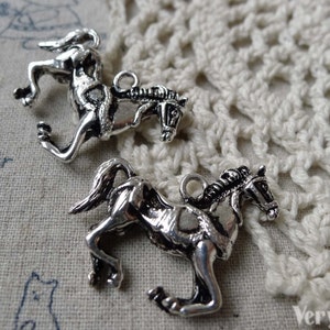 May include: Two silver-toned horse charms. The charms are detailed with a running horse design, featuring a mane, tail, and a loop for attaching to jewelry. The charms are on a textured, off-white surface.