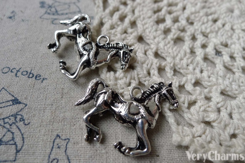 May include: Two silver-toned horse charms. The charms are detailed with a running horse design, featuring a mane, tail, and a loop for attaching to jewelry. The charms are on a textured, off-white surface.