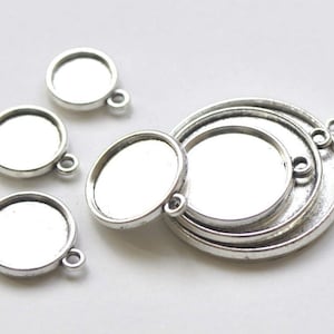 May include: A set of five silver-toned metal pendant trays for jewelry making. The trays are round and have a smooth, reflective surface. They are perfect for adding a touch of elegance to your jewelry designs.