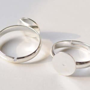 May include: Two silver adjustable rings with round, flat, silver-colored bezels. The bezels are designed to hold a cabochon or other flat-backed embellishment.