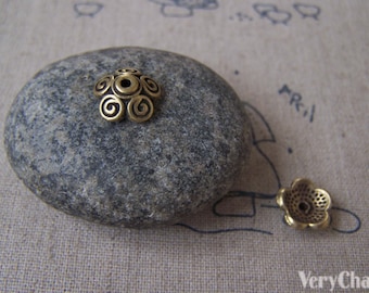 Puede incluir: Dos tapas de cuentas de metal de color latón antiguo. Una tiene forma de cúpula con diseños en espiral, y la otra tiene forma de flor. Las tapas de las cuentas están sobre una piedra gris, con un fondo neutro.